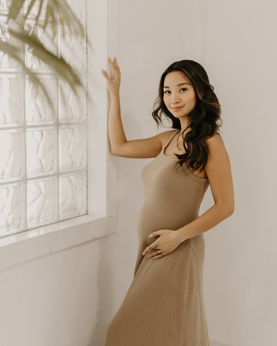 Nika in a full-length taupe dress beside a softly lit glass wall.