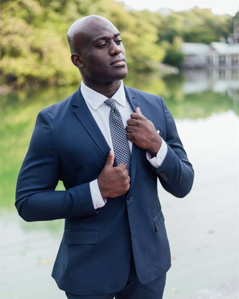Emeka adjusting his navy suit beside the water.